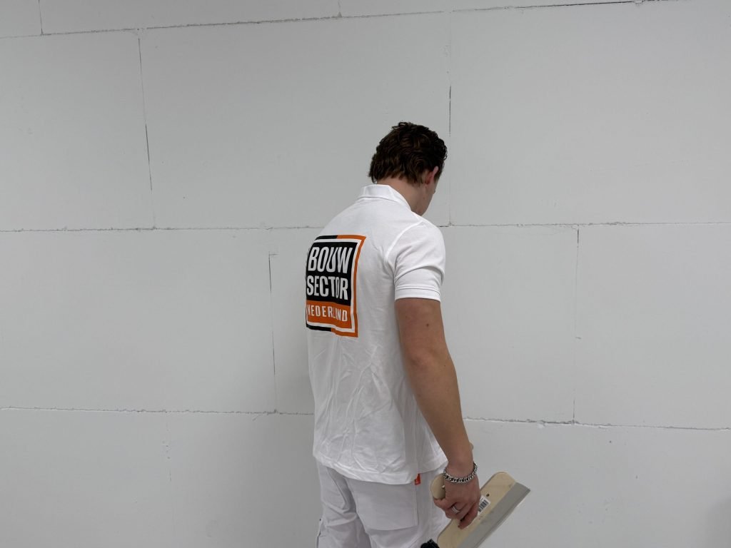 Vakman van Bouwsector Nederland bekijkt een kale wand met zichtbare voegen, met een spaan in de hand